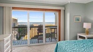 A bedroom with a balcony overlooking the beach, a bed, a chair, and a small table with a lamp. - Trip's OBX: Oceanfront Townhome, Pool, Tennis (Nags Head)