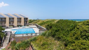 A pool area with a view of the ocean, surrounded by greenery and a building with balconies. - Trip's OBX: Oceanfront Townhome, Pool, Tennis (Nags Head)