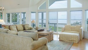 A spacious living room with a large window overlooking a body of water, a comfortable sofa set, and a dining area with a table and chairs. - Links End: Soundfront, Hot tub, Shared pool, Golf (Nags Head)