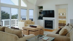 A spacious living room with a fireplace, large windows, and a balcony. - Links End: Soundfront, Hot tub, Shared pool, Golf (Nags Head)