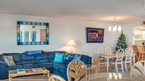 A living room with a blue sofa, a coffee table, wicker chairs, a dining table with chairs, a chandelier, and a Christmas tree.