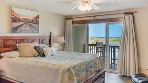 Ein Schlafzimmer mit Bett, Deckenventilator, einem Gemälde und Blick auf den Strand durch eine Schiebetür.