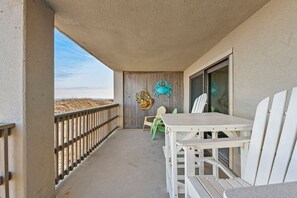 A balcony with a table and chairs, a decorative crab, and a view of the beach. - Heron Cove 204S: Oceanfront Condo, Shared Pool (Nags Head)