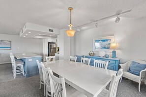 A modern kitchen with white cabinetry, a central island with seating, and a dining area with white chairs. - Heron Cove 204S: Oceanfront Condo, Shared Pool (Nags Head)