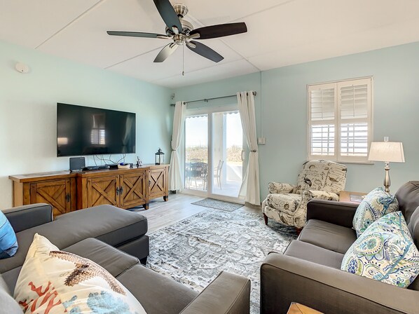 A living room with a ceiling fan, a flat-screen TV, a wooden cabinet, a patterned armchair, and a sofa with decorative pillows.