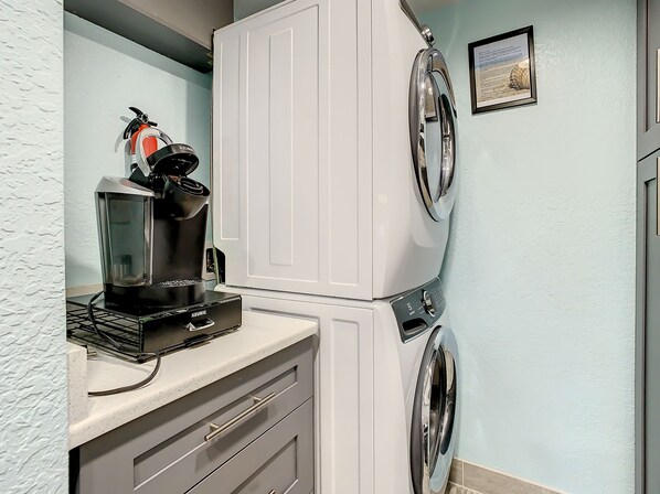 A compact laundry area with a front-loading washer and dryer, a coffee maker, and a small shelf.