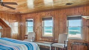 Habitación de cabaña de madera con una cama, dos sillas, una mesita y ventanas amplias con vista al océano.