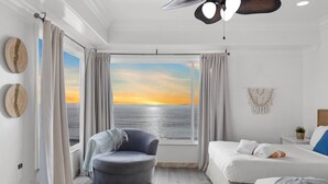A room with a large window offering a view of the sea at sunset, a bed, a chair, and a ceiling fan.