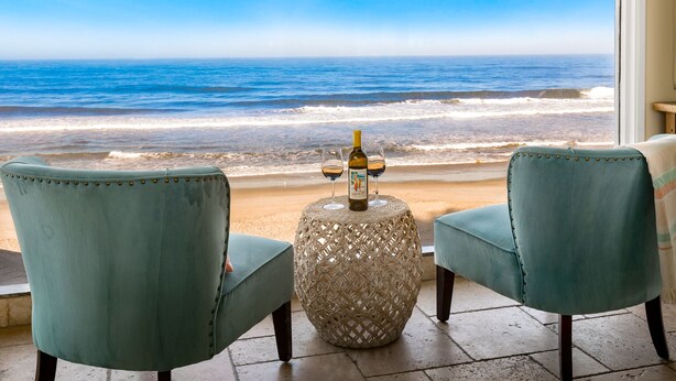 Ein Zimmer mit zwei türkisfarbenen Sesseln, die auf einen Strand Blick bieten, einem gewebten Tisch mit einer Flasche und Gläsern sowie einem großen Fenster mit Meerblick.