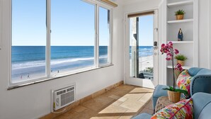 Ein Zimmer mit großem Fenster zum Strand, einem kleinen Balkon und einer weißen Regalanlage mit Pflanzen.