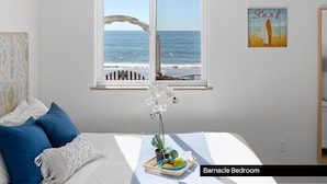 Ein Schlafzimmer mit großem Fenster mit Blick auf den Strand, einem Bett mit blauen Kissen, einer Pflanze im Fensterbrett und einem 'Surf'-Poster an der Wand.
