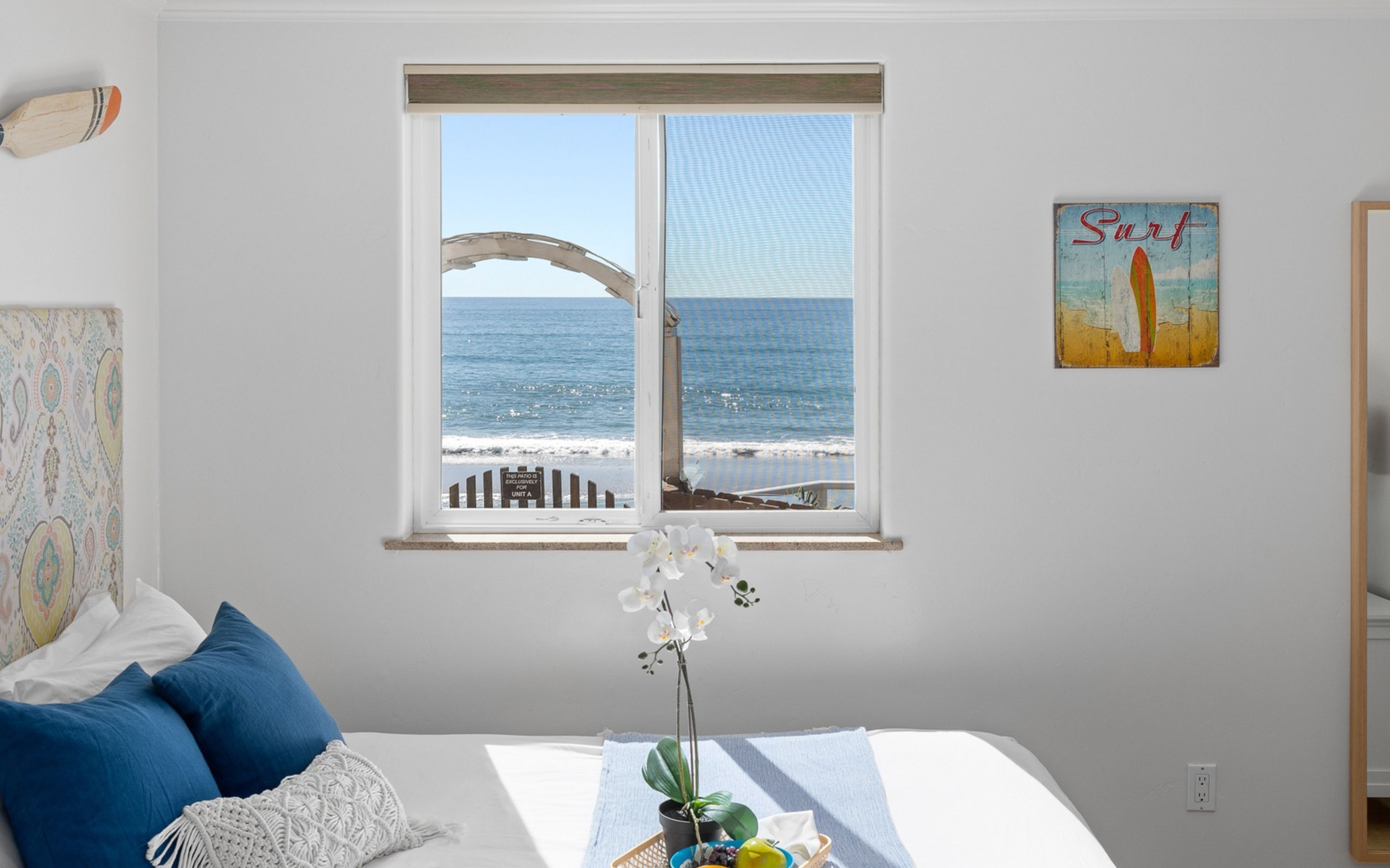 A bedroom with a large window overlooking the beach, a bed with blue pillows, a plant on the windowsill, and a 'Surf' poster on the wall.