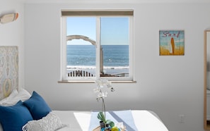 Une chambre avec une grande fenêtre donnant sur la plage, un lit avec des oreillers bleus, une plante sur le rebord de la fenêtre et une affiche sur le thème du surf accrochée au mur.