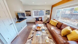A living room with a brown leather sofa, a coffee table, a television on a stand, and a window with a view of the outside. - Moab Flats - 4, All-Star Downtown Location, Pet Friendly (Moab)