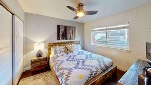 Un dormitorio con cama, mesita de noche, lámpara, ventilador de techo, ventana y una mesa de madera.