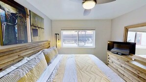 A bedroom with a bed, a television on a wooden dresser, a window with a view of the outside, and artwork on the wall.