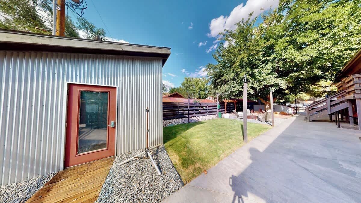 A corrugated metal building with a red door, a paved walkway, and a grassy area with trees.