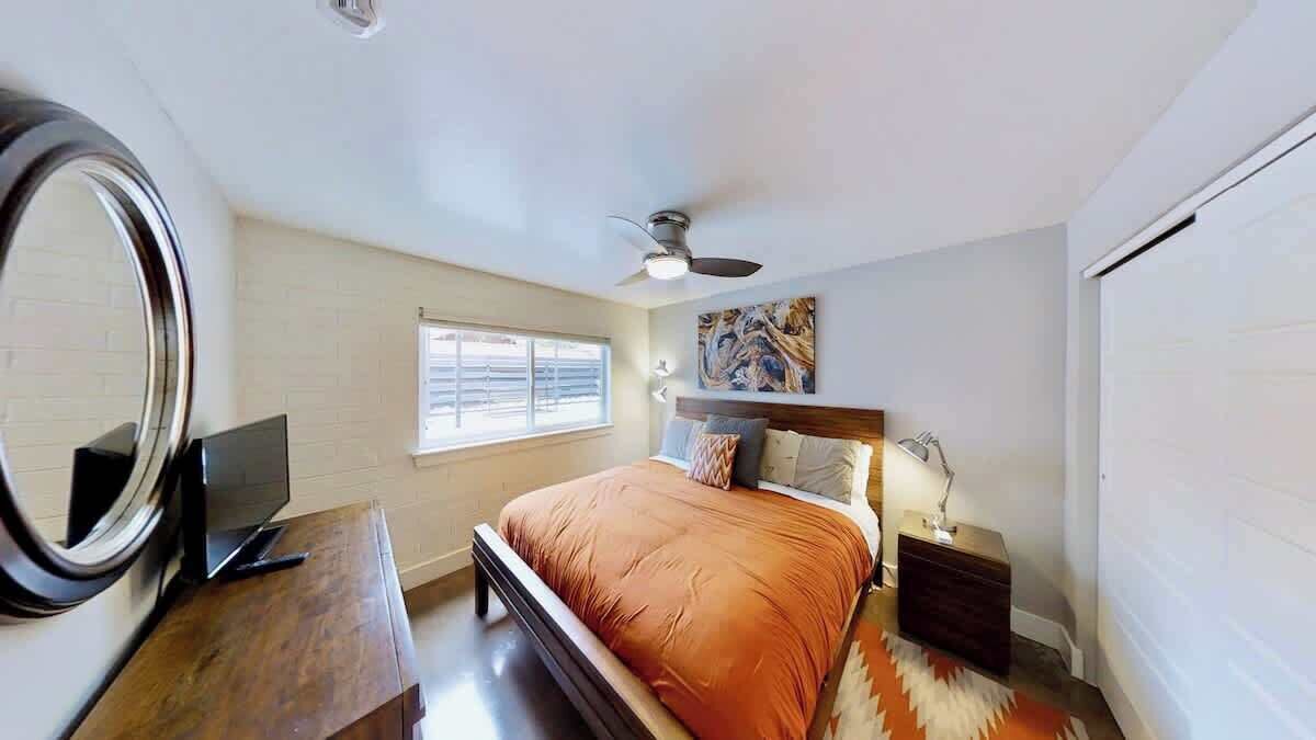 A bedroom with a bed, a desk, a mirror, a ceiling fan, and a window.
