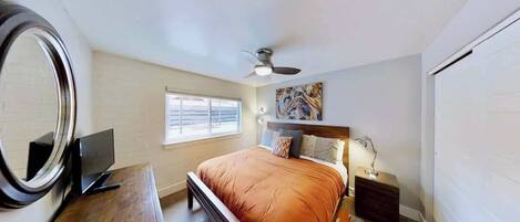 A bedroom with a bed, a desk, a mirror, a ceiling fan, and a window.