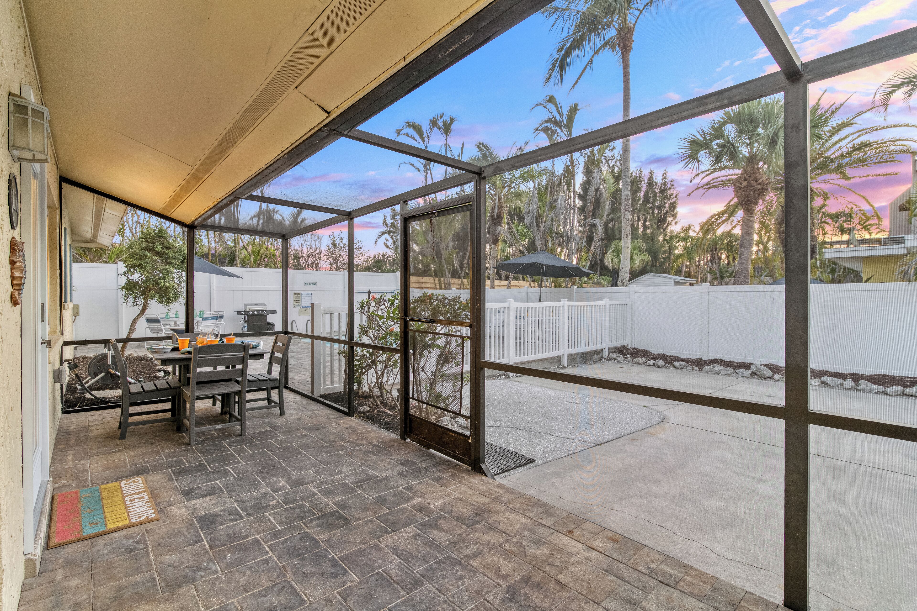 A covered patio with a table and chairs, a view of palm trees and a white fence, and a clear sky.