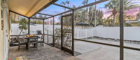 A covered patio with a table and chairs, a view of palm trees and a white fence, and a clear sky.