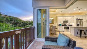 A balcony with a wooden railing, a wicker sofa with cushions, and a sliding glass door leading to a kitchen with white cabinets, a microwave, and a stainless steel refrigerator.