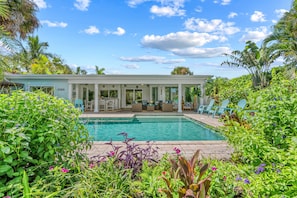 A poolside area with a covered seating area, a pool, and lush greenery.