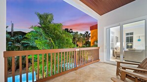 A balcony with a wooden railing, a pool, and a view of lush greenery and a sunset.