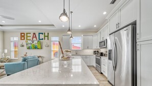 A modern kitchen with white cabinetry, a large island, stainless steel appliances, and a 'BEACH' sign on the wall.