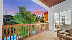 A balcony with a wooden railing, a pool, and a view of lush greenery and a sunset. - Casa Mahalo | Pet Friendly, Ping Pong Table, Private Heated Pool, Steps to Beach! (Siesta Key)