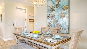 Comedor con mesa de madera dispuesta para cuatro personas, con platos y vasos azul claro, un jarrón de flores amarillas y un cuadro abstracto grande en la pared.