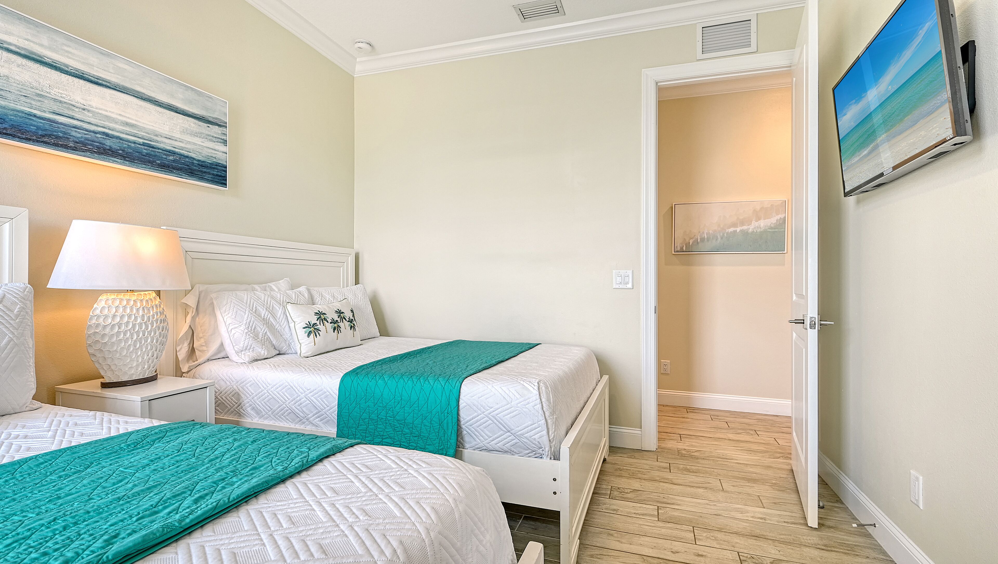 Un dormitorio con dos camas, una mesita de noche con lámpara, un cuadro en la pared y una puerta que da a otra habitación.