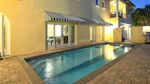 A swimming pool with a brick border, a building with a balcony, and a striped awning. - 🦋 Casa De Mariposa | Heated Pool, Gulf Views, Dual-Villa Duplex, Walk to Siesta Key Beach (Siesta Key)