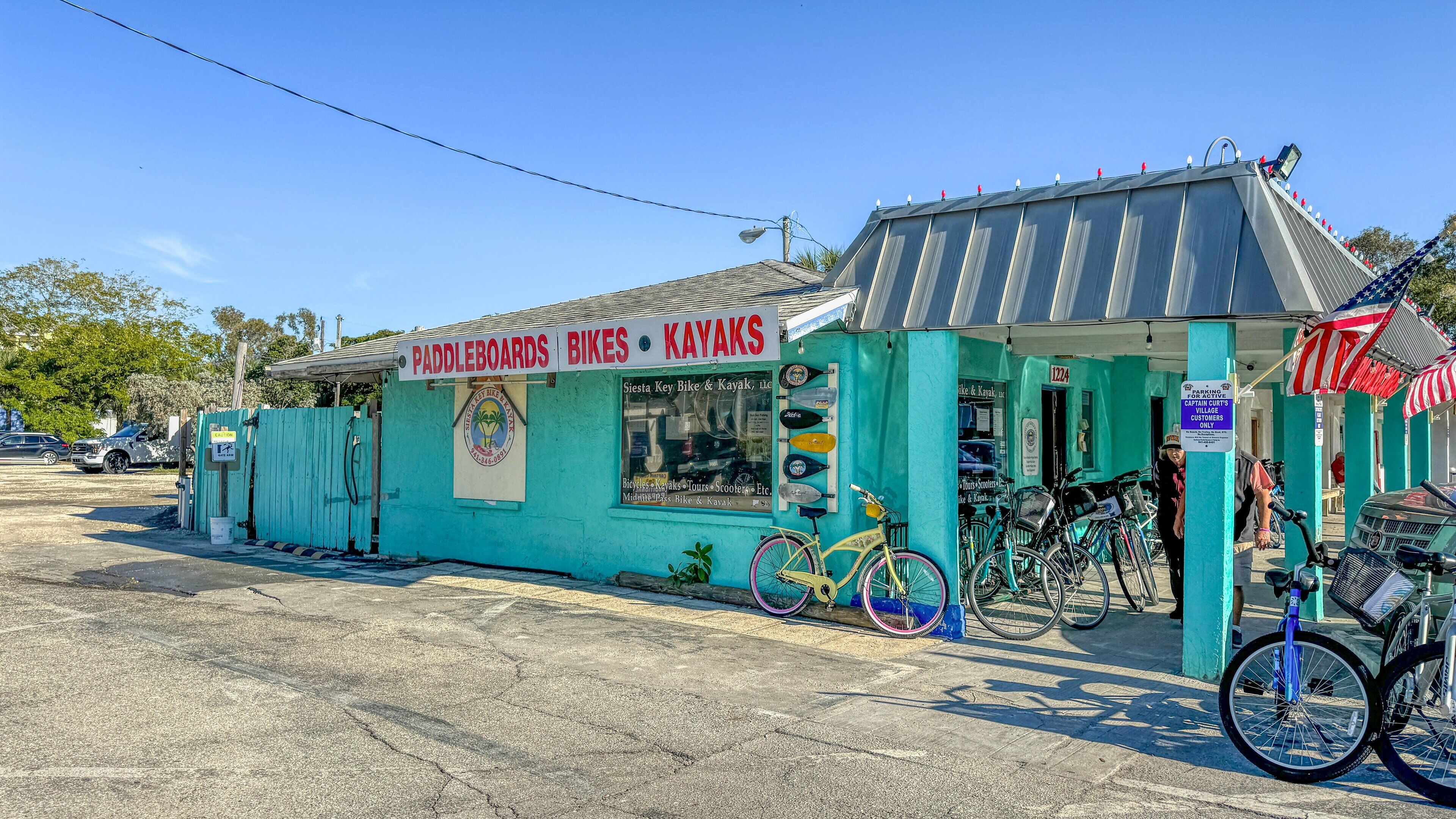 A small outdoor kiosk with a sign advertising paddleboards, bikes, and kayaks. There are several bicycles parked in front, and the kiosk has a teal exterior with a corrugated metal roof.