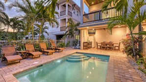 A pool area with lounge chairs and a dining table, surrounded by palm trees and a building with balconies.
