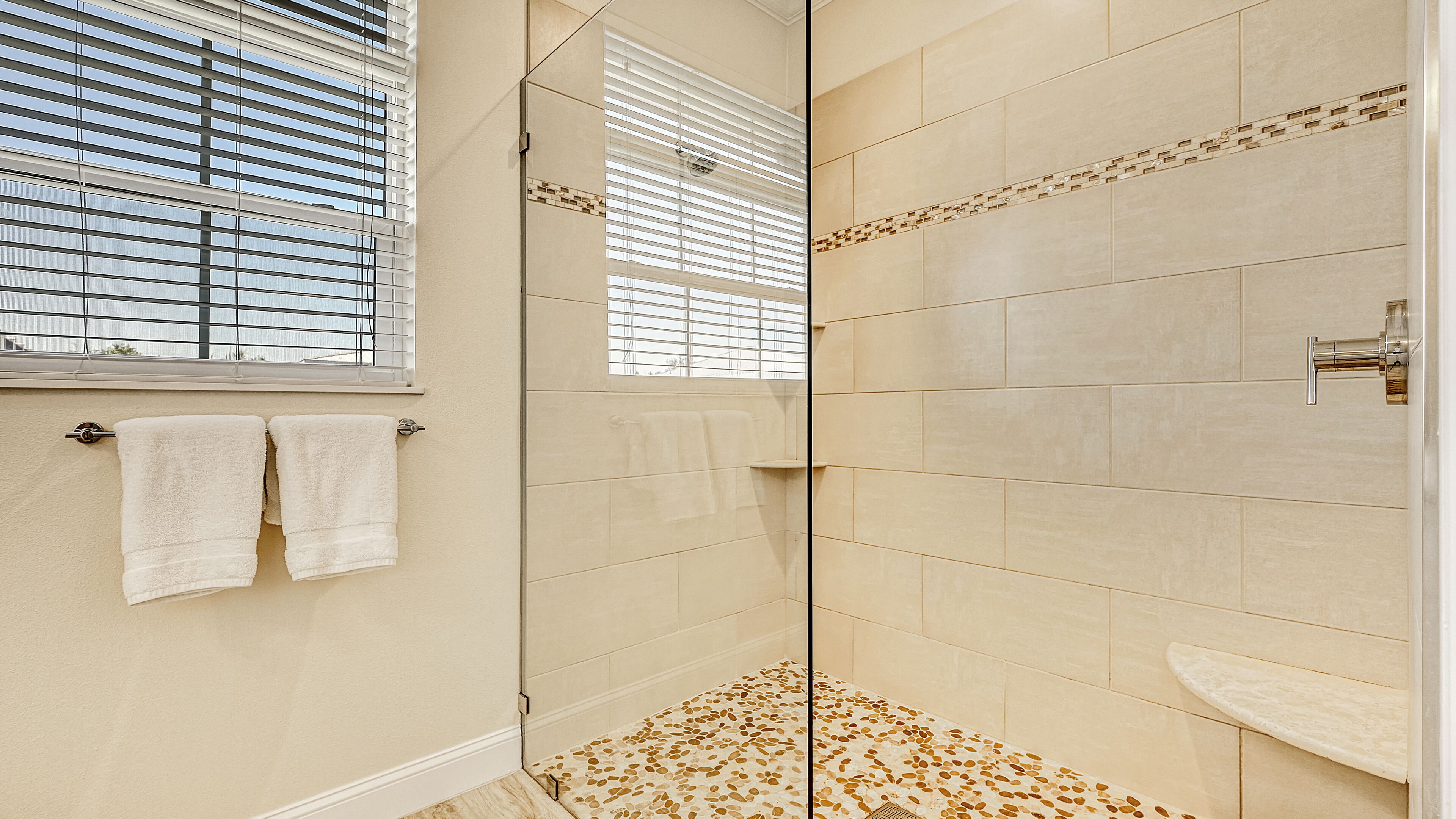 A modern bathroom with a glass-enclosed shower, beige tiles, and a window with blinds.