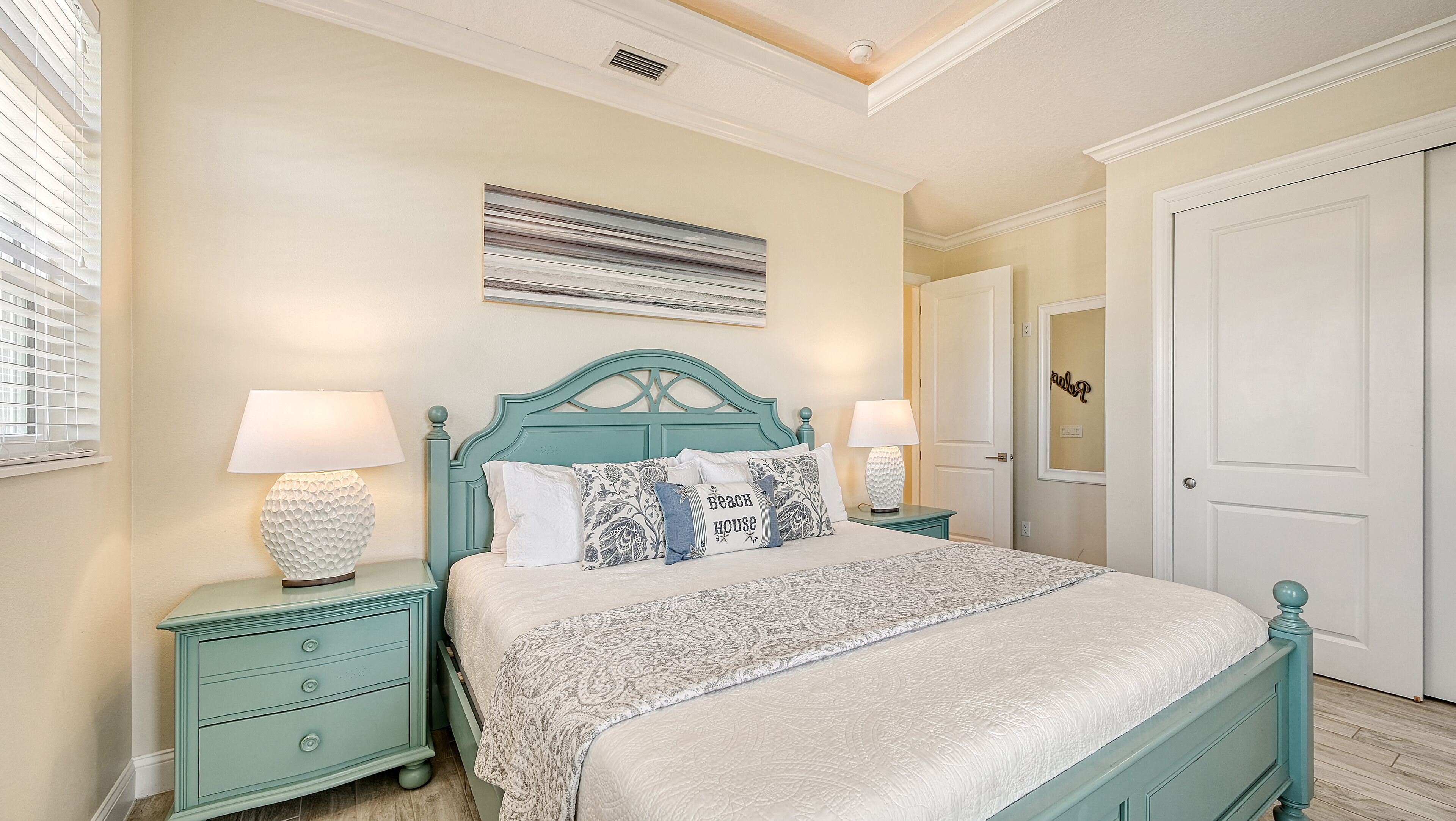 A bedroom with a teal-colored bed, two bedside tables with lamps, a patterned rug, and a door with a mirror.