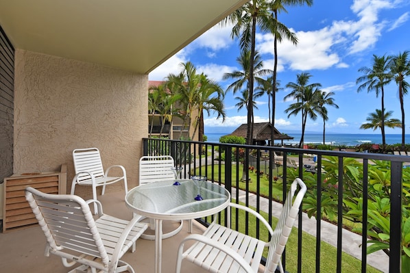 A balcony with white metal chairs and a glass table, overlooking a beach with palm trees and a view of the ocean.