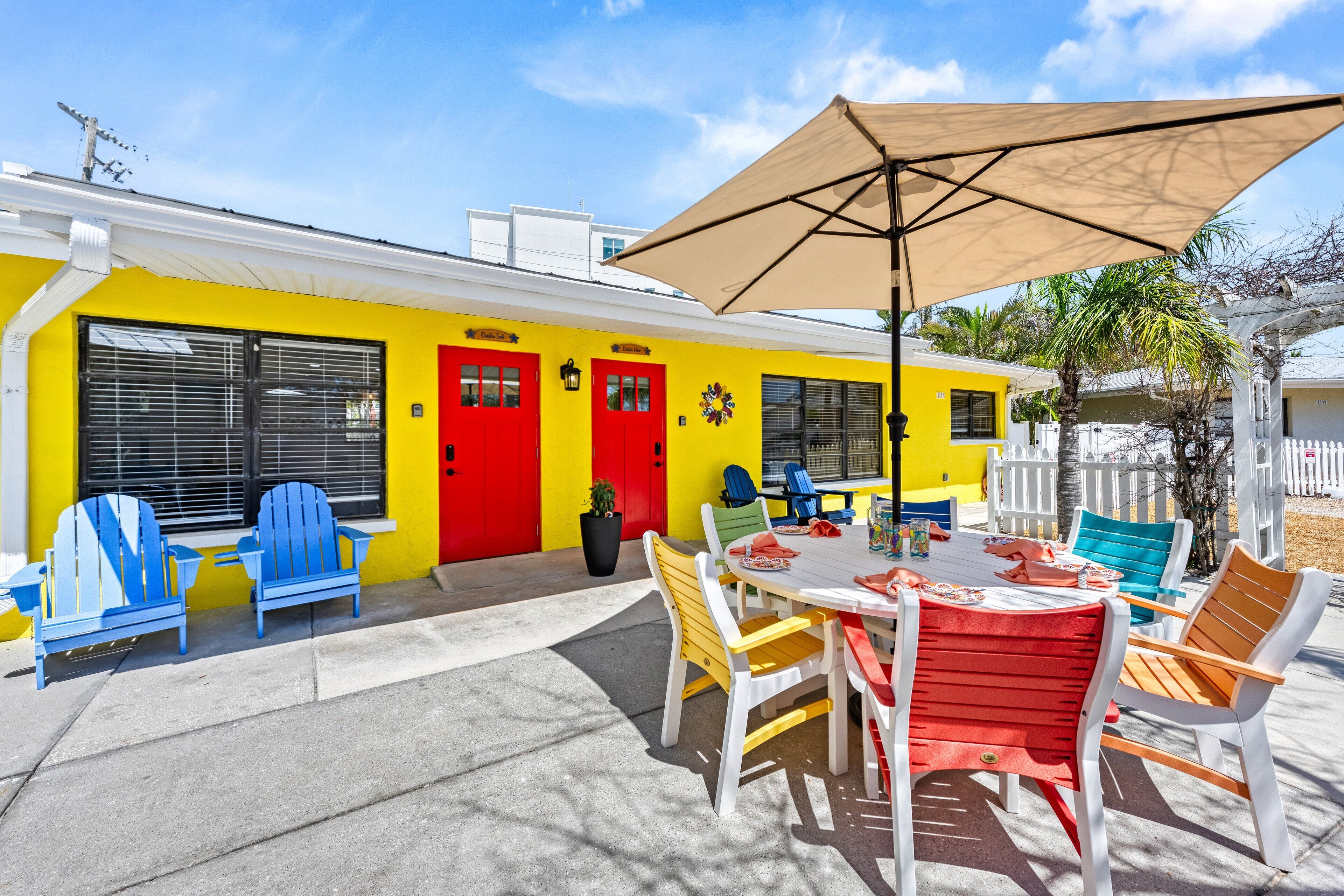 A patio with a table and chairs, a yellow building with red doors, and a white fence in the background.