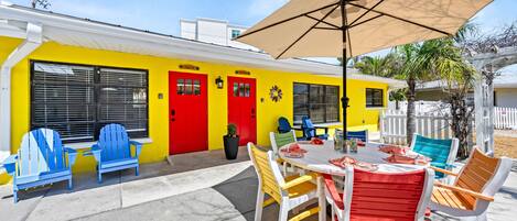 A patio with a table and chairs, a yellow building with red doors, and a white fence in the background.