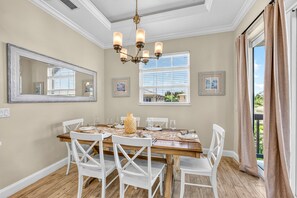 Uma sala de jantar com mesa de madeira posta para quatro pessoas, cadeiras brancas, um lustre e quadros nas paredes.