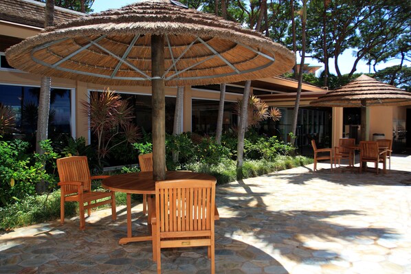 An outdoor patio with wooden furniture, stone flooring, and thatched umbrellas. - Outstanding Oceanfront Views - Mahana 1017 (Lahaina)