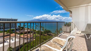 A balcony with a view of the ocean and a mountain, white lounge chairs, and a building in the background.