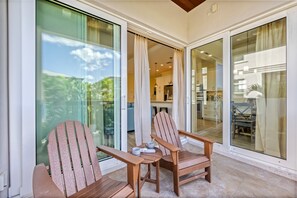 A balcony with two wooden chairs and a table, a sliding glass door leading to a kitchen, and a view of greenery outside.