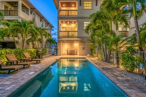A swimming pool with lounge chairs and a barbecue grill, surrounded by palm trees and a building with multiple balconies.