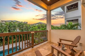 A balcony with a view of a swimming pool and surrounding greenery, wooden furniture, and a ceiling fan.