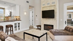 Un salon moderne doté d’une cheminée, d’un téléviseur et d’un coin cuisine avec des meubles blancs et un pare-feu en pierre.