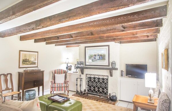 Una sala de estar con vigas de madera a la vista, una chimenea, un televisor y un reposapiés verde.