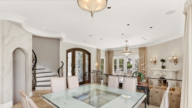 Un comedor espacioso con una mesa y sillas de vidrio, una escalera con barandillas ornamentales y una chimenea con apliques decorativos.