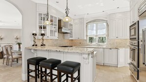 Una cocina amplia con armarios blancos, una isla grande con taburetes de bar, electrodomésticos de acero inoxidable e iluminación colgante.